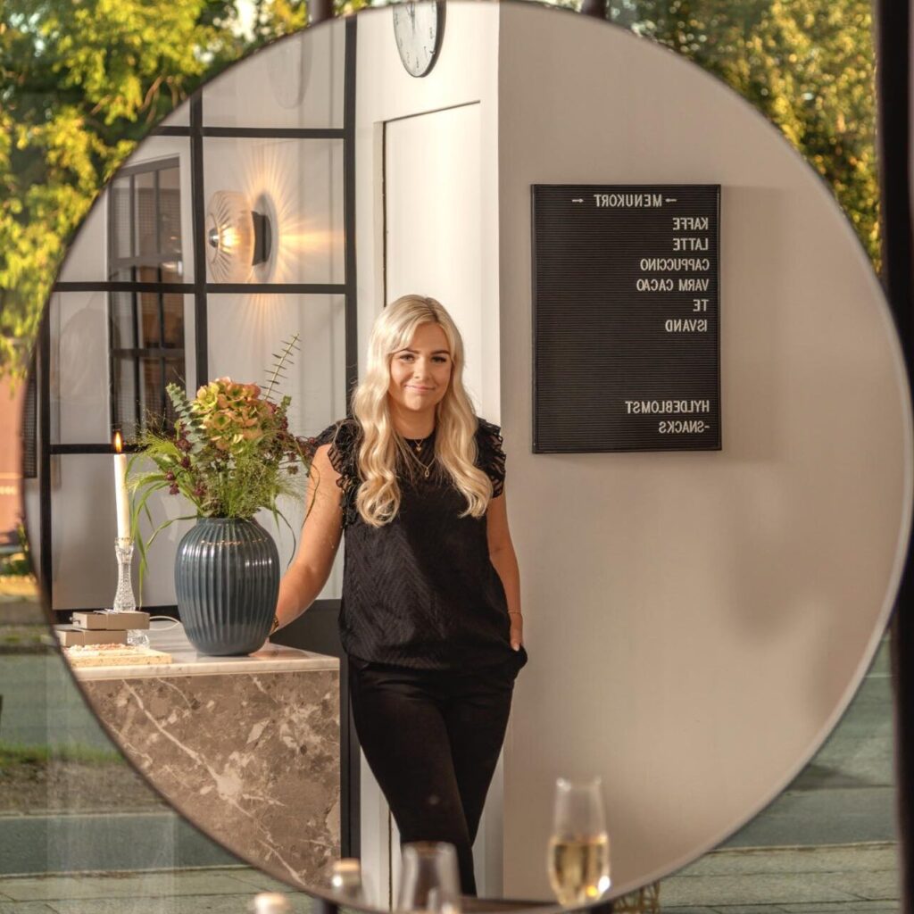 Frisør Pia Rasmussen står smilende i sin moderne salon i Risskov, spejlet reflekterer hyggelig atmosfære med champagne og snacks